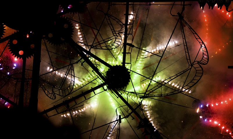 The intricate gears and framework of catherine wheels silhouetted by the flare and spark of another catherine wheel behind it.